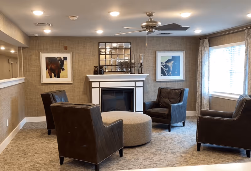A cozy seating area in a senior living facility with four dark leather armchairs arranged around a large round beige ottoman. The room features a white fireplace with decorative items on the mantel, a large window with curtains allowing natural light, and two framed pictures on the wall. The ceiling has recessed lighting and a modern ceiling fan.