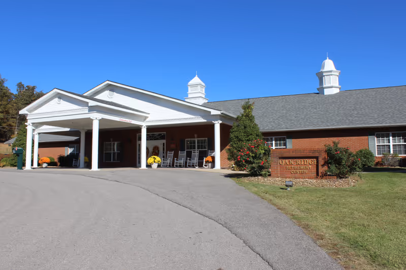Front exterior view of Oak Ridge Retirement Center building with a covered entrance supported by white columns, rocking chairs on the porch, and a sign in front surrounded by bushes and flowers under a clear blue sky.