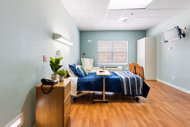 A hospital-style bedroom in a senior living center with a bed, dresser, and plants.