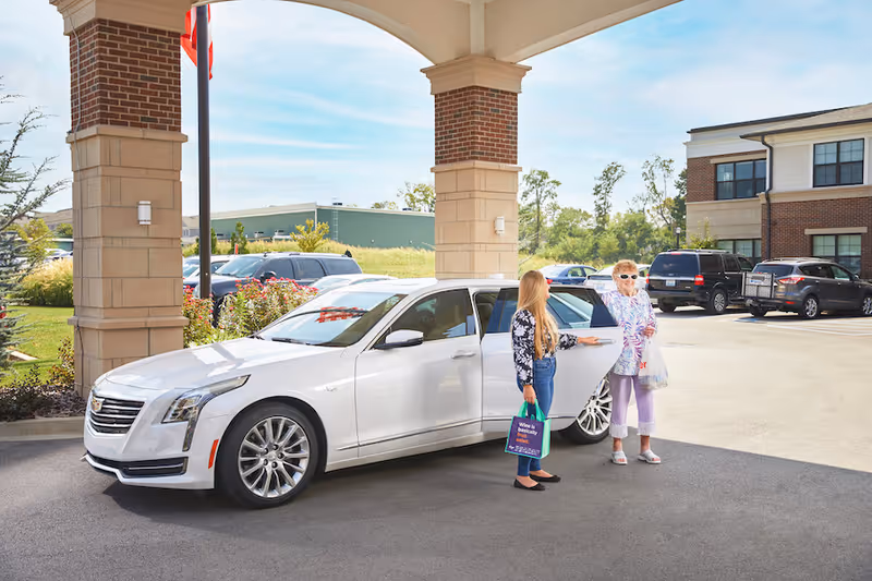 A young woman holding a reusable shopping bag opens the door of a white luxury car for an elderly woman standing outside near the entrance of a senior living facility. The elderly woman is wearing sunglasses and holding a plastic bag. Several parked cars and a two-story brick building are visible in the background under a clear sky.