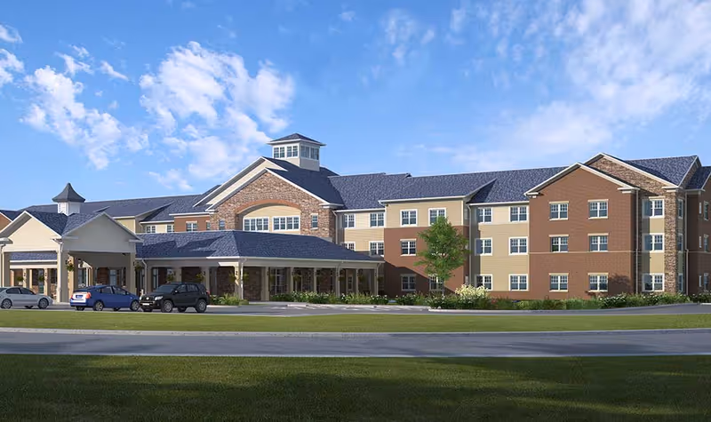 Exterior view of a large senior living facility building with multiple stories, a covered entrance, several windows, and a few cars parked in front under a partly cloudy blue sky.