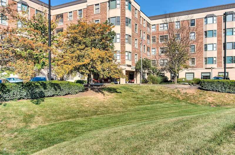 Exterior view of The Lafayette senior living facility showing a multi-story brick building with several windows, surrounded by green grass, trees with autumn foliage, and shrubs under a clear blue sky.