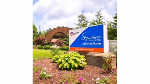 Outdoor view of the Signature HealthCARE at Jefferson Manor Rehab & Wellness Center sign in landscaped grounds with the facility building visible behind it.