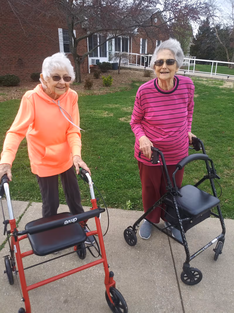 Two elderly women standing outside on a sidewalk in front of a brick building with green grass and trees. Both women are using walkers and wearing glasses. One woman is wearing an orange hoodie and dark pants, while the other is wearing a pink and purple striped long-sleeve shirt and maroon pants. They are smiling and appear to be enjoying the outdoor setting.