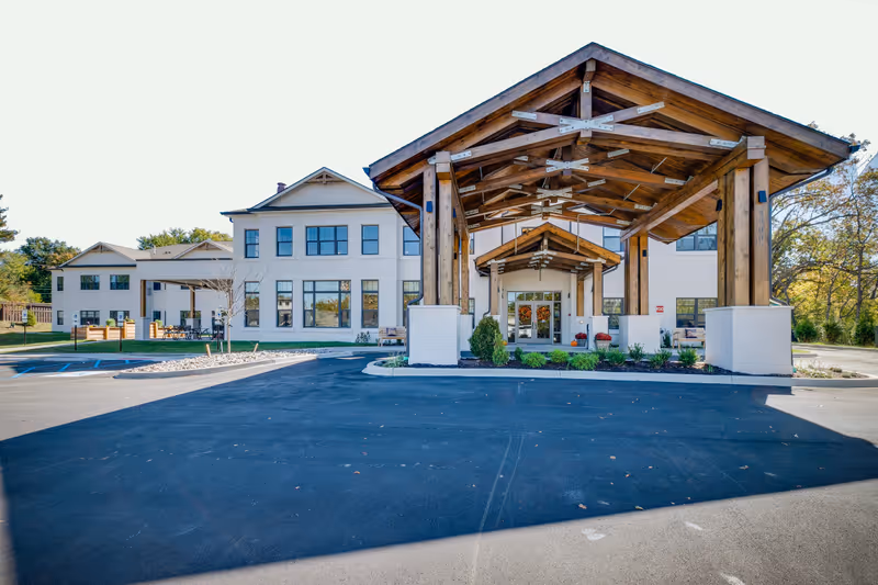 Front entrance of a two-story senior living facility with a large wooden covered porte-cochère and a paved parking area.