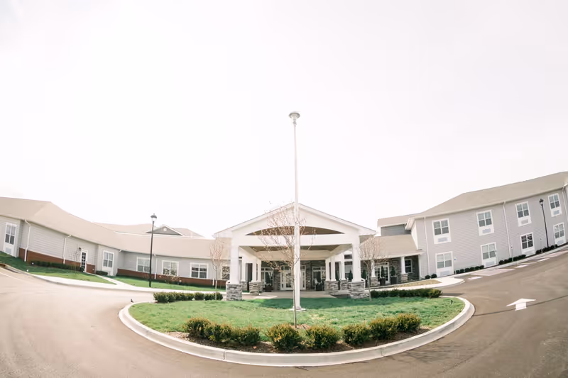 Front exterior view of Bluegrass Senior Living facility showing a large entrance with a covered drop-off area, surrounded by a circular driveway and landscaped greenery.