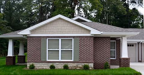 Front exterior of a single-story brick building with a central window, covered porches, and an attached garage surrounded by grass and trees.