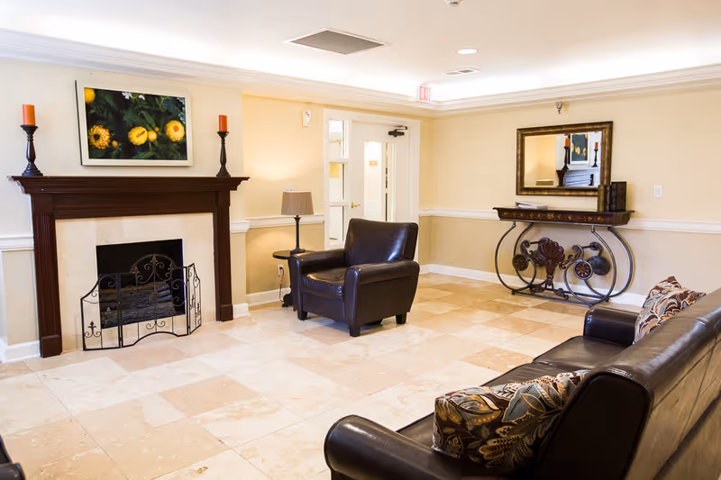 A cozy living room area with beige tiled flooring, cream-colored walls, and a dark wooden fireplace mantel with a decorative fire screen. Above the mantel is a framed picture of yellow flowers. Two dark leather armchairs and a matching sofa with patterned cushions are arranged in the room. A decorative console table with a large mirror above it is against the wall. The room is well-lit with recessed ceiling lights and a table lamp near the armchair. A door with glass panels is visible in the background.