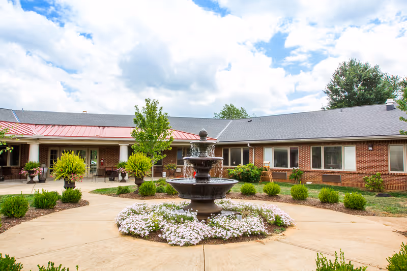 A landscaped courtyard with a tiered fountain in front of a single-story brick senior living facility.