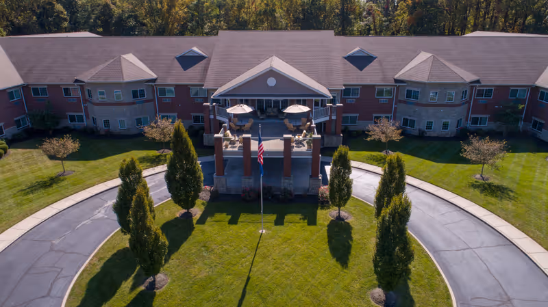 Aerial view of the front exterior of Magnolia Springs East Louisville, showing a large two-story building with a covered entrance, outdoor seating with umbrellas, a circular driveway, neatly trimmed grass, and several trees.
