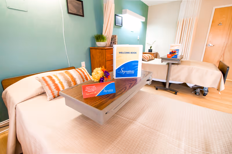 A bright and clean room with two hospital-style beds, each with beige bedding and decorative pillows. Each bed has an overbed table with a welcome book and a small card on it. The walls are painted green and light beige, and there is a wooden dresser with a plant on top. The room has a wooden door and a light-colored floor.