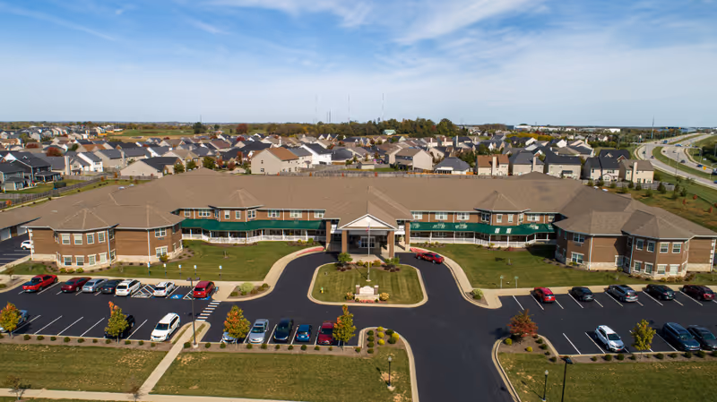 Aerial view of Magnolia Springs Lexington, a large senior living facility with a U-shaped building surrounded by parking lots and green lawns. The building has a brown roof and beige walls with green awnings over the windows. The facility is located in a suburban area with many houses and a highway visible in the background under a partly cloudy sky.