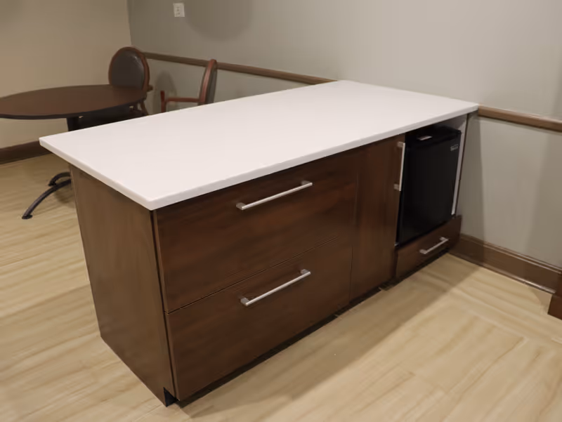 A wooden kitchen island with a white countertop, two drawers, a cabinet, and a small black refrigerator. In the background, there is a round table with two chairs against a beige wall.