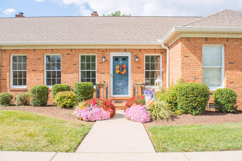 Front exterior view of a single-story brick building with a blue door decorated with a fall wreath. The entrance is flanked by two black lantern-style lights and surrounded by neatly trimmed bushes and colorful flower beds. A concrete pathway leads up to the door under a clear blue sky.
