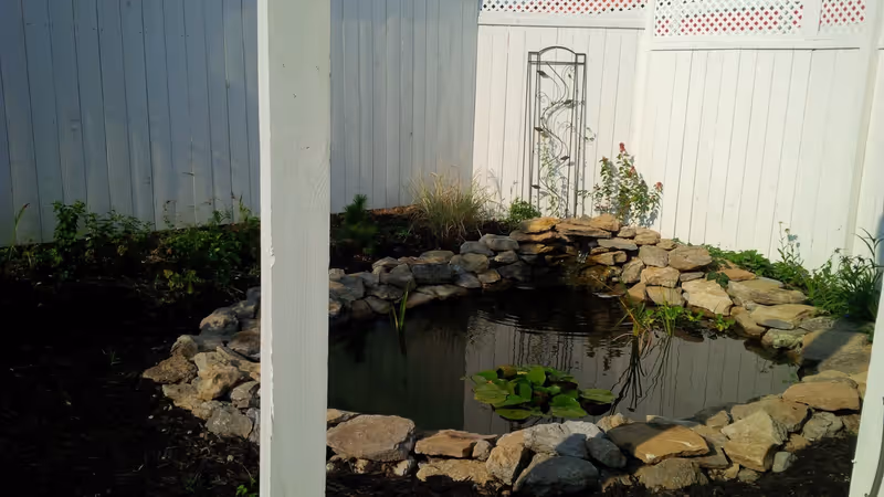 A small garden pond surrounded by rocks with water lilies floating on the surface, set against a white wooden fence with some plants and a metal trellis in the background.