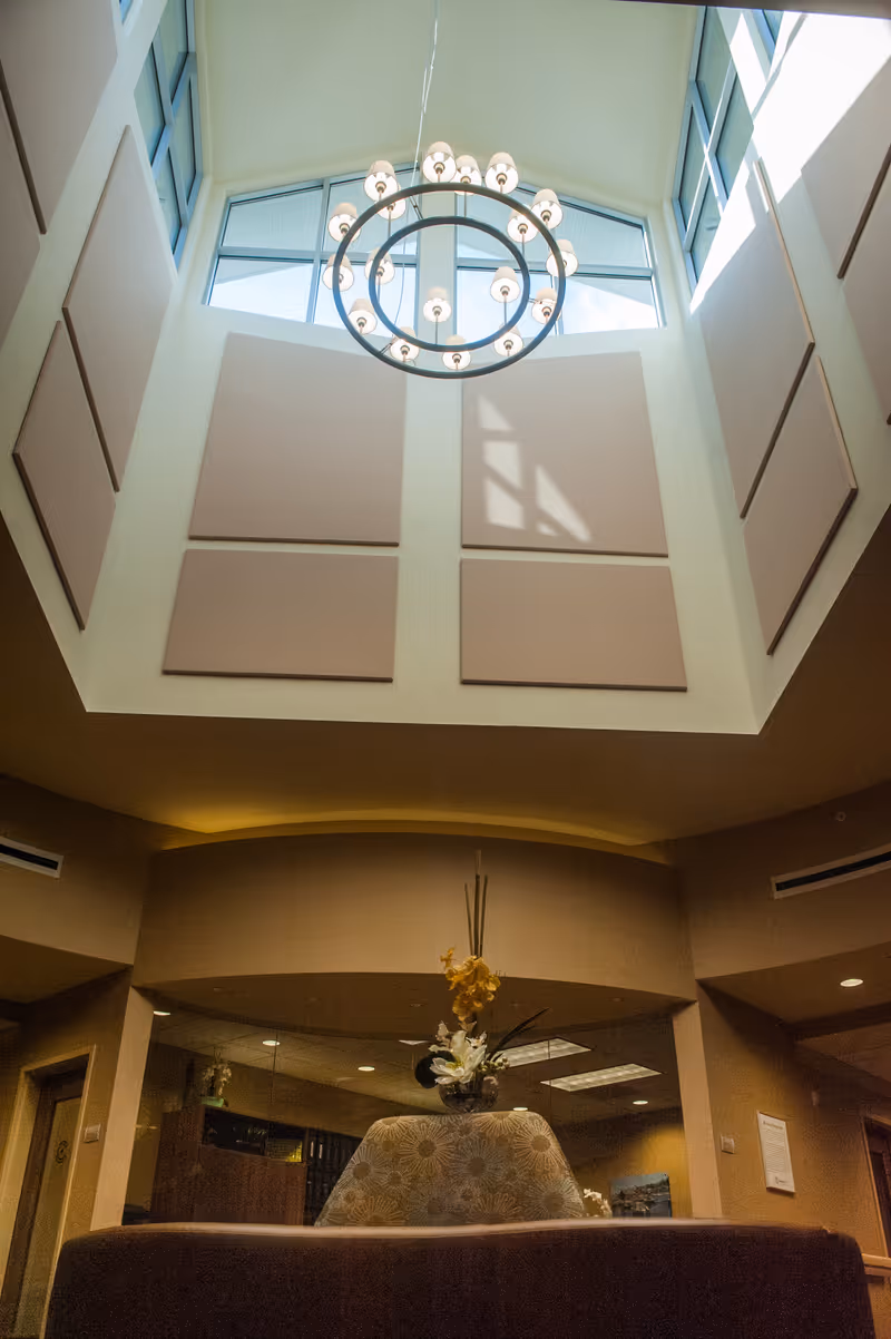 Interior view of a facility lobby or common area with a high ceiling, large windows near the roof, a modern circular chandelier with multiple lights, and a floral arrangement on a patterned piece of furniture in the foreground.