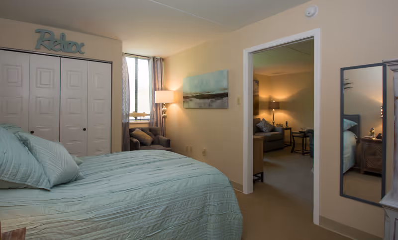 A cozy bedroom with a neatly made bed covered in light blue bedding. There is a window with curtains and a small armchair next to it. Above the closet doors, a decorative sign reads 'Relax'. The room opens into a softly lit living area with a sofa, side table, and lamp. A full-length mirror is mounted on the wall near the doorway.