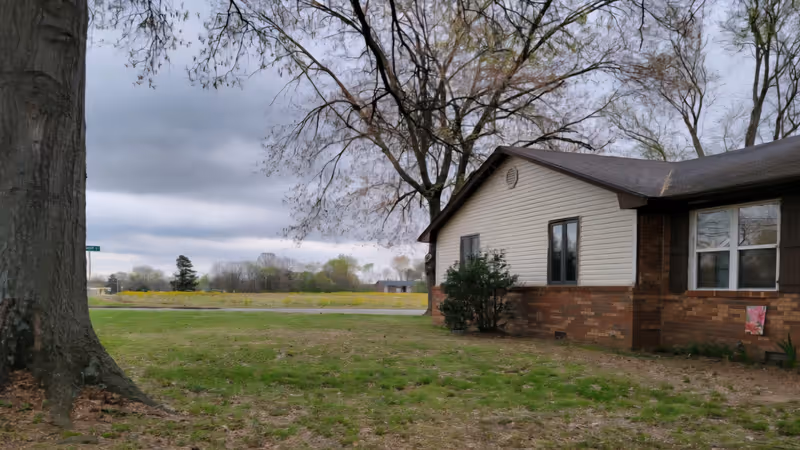 A single-story brick and beige siding building sits beside a grassy yard with trees.