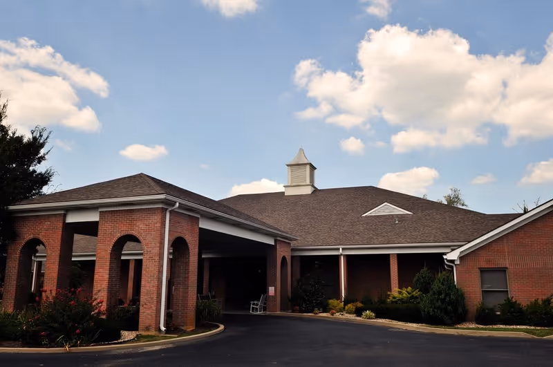 Exterior view of a single-story brick building with a covered entrance supported by brick columns. The building has a dark shingled roof with a small cupola on top. There are shrubs and landscaping around the building, and the sky is partly cloudy.