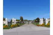 Wide paved driveway entrance flanked by white pillars and landscaped bushes leading into a residential or senior living community under a clear blue sky.