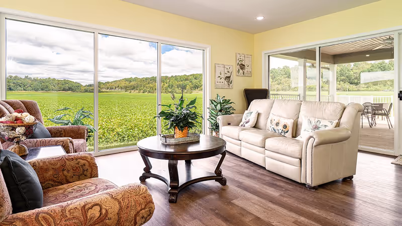 A bright living room with large sliding glass doors offering a view of a green field and trees outside. The room features a beige leather sofa with floral pillows, two patterned armchairs, a round wooden coffee table with a potted plant, and light yellow walls with botanical artwork. Outside on the patio, there is a table with chairs.