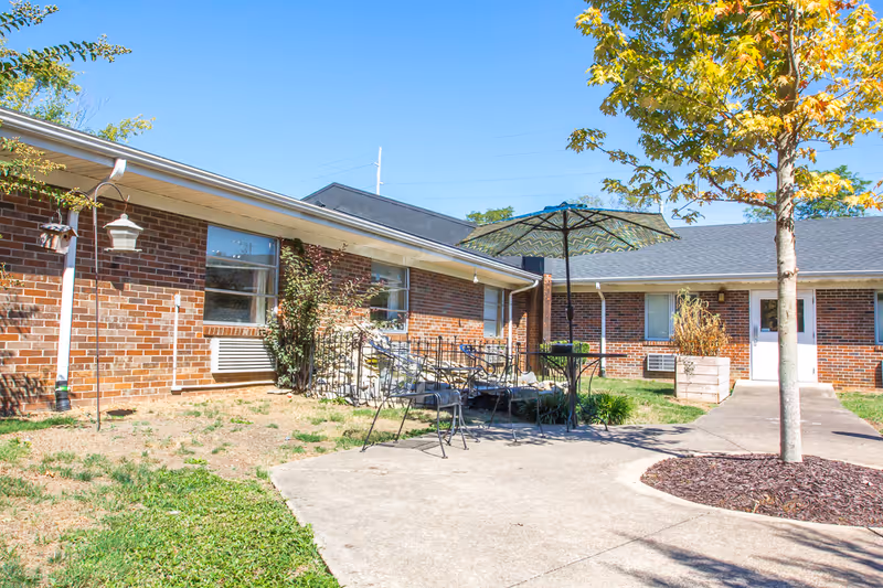 Courtyard outside a single-story brick rehab center with patio tables, an umbrella, a walkway and a tree.