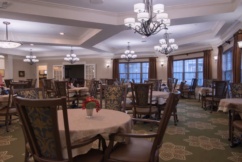 A spacious dining room with multiple round tables covered with beige tablecloths, each table surrounded by wooden chairs with patterned upholstery. The room is well-lit with several chandeliers hanging from the ceiling and large windows with brown curtains allowing natural light to enter. The carpet has a floral pattern, and small flower arrangements are placed on the tables.