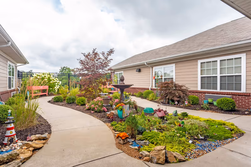 A landscaped courtyard with a winding concrete path, colorful flower beds, a central fountain and benches between single-story buildings.