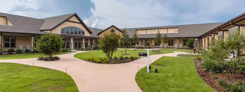 Outdoor courtyard area of The Legacy at English Station featuring a paved walkway, green lawns, trees, benches, and a mailbox, surrounded by a single-story building with large windows and a sloped roof under a partly cloudy sky.
