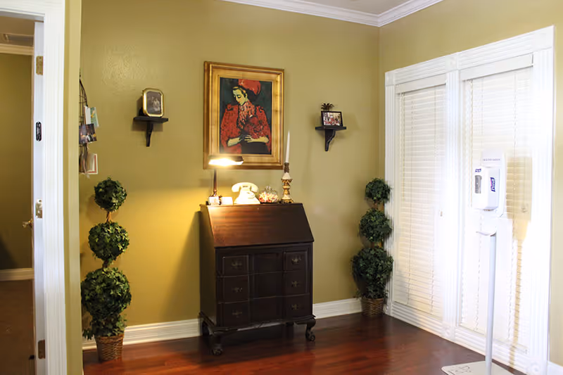 A small, furnished interior corner with a dark wood cabinet, painting, lamp, and topiary.