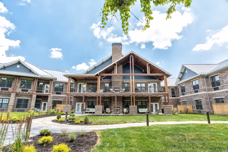 Exterior view of a two-story senior living facility building with a large covered balcony and patio area. The building features brick walls, multiple windows, and a metal roof. There is a well-maintained lawn with landscaped plants and a curved walking path in the foreground under a partly cloudy blue sky.