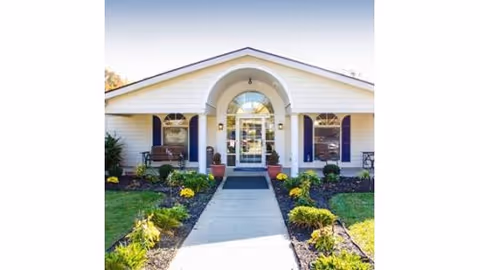 Front exterior view of a single-story building with white siding and a central entrance featuring an arched window above the door. There are two windows on either side of the entrance with dark shutters, a bench on the left side, and landscaped flower beds with green shrubs and yellow flowers along the walkway leading to the door.