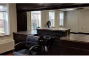 Interior view of a reception or front desk area with dark wood counters and a black office chair. There are large windows with white blinds allowing natural light into the room. A small decorative item is placed on the counter.