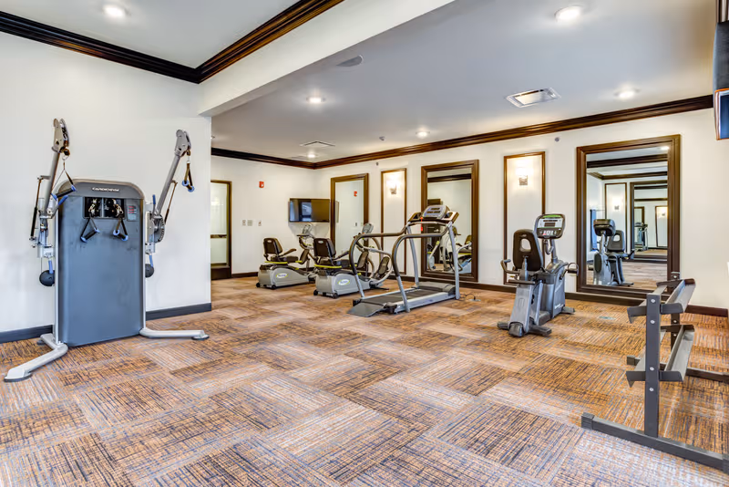 Indoor senior living fitness center with cardio machines and weight equipment.