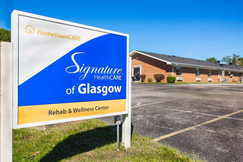 Outdoor view of Signature HealthCARE of Glasgow Rehab & Wellness Center sign with a brick building and parking lot in the background under a clear blue sky.