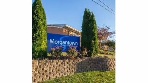A blue sign reading "Morgantown" at the facility entrance with shrubs, a stone retaining wall and tall evergreen trees.