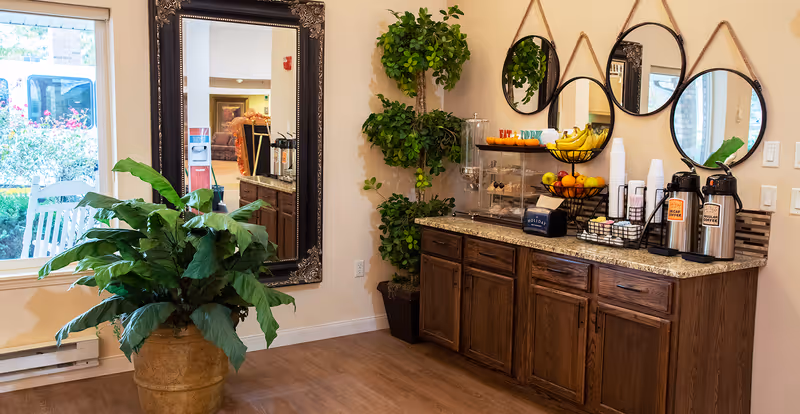 A cozy refreshment area in a senior living facility with a wooden cabinet topped with granite, holding coffee dispensers labeled decaf and regular, a basket of bananas and other fruits, a container with snacks, cups, and sweeteners. Above the cabinet are four round mirrors hanging on the wall. To the left, a large potted plant sits on the wooden floor near a window with a view of a white rocking chair and greenery outside. A tall green plant is placed between the cabinet and a large ornate framed mirror reflecting part of the room.