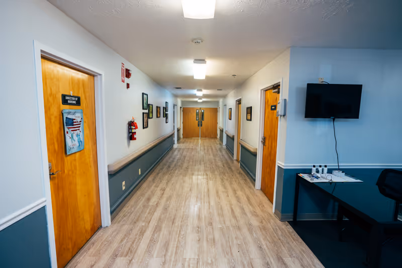 A well-lit hallway in a senior living facility with light wood flooring and white walls with blue wainscoting. Several wooden doors line the hallway, including one labeled 'Director of Nursing.' There are framed pictures on the walls, a fire extinguisher, and a wall-mounted TV above a desk with papers and glasses on it.