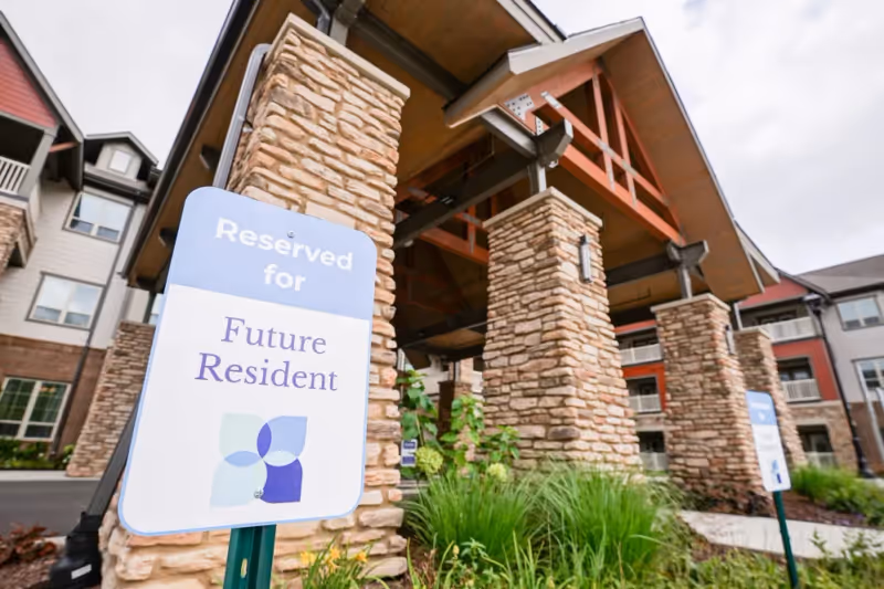Entrance area of a senior living facility with stone pillars and a covered driveway. A sign in the foreground reads 'Reserved for Future Resident'. The building has multiple floors with balconies and a mix of stone and siding exterior.