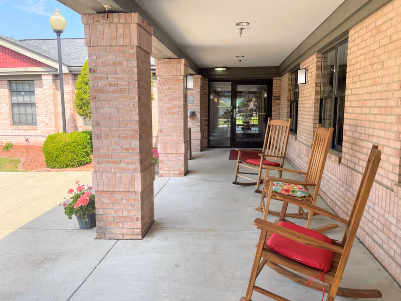 Covered outdoor porch area with three wooden rocking chairs, each with a cushion, facing outward. The porch has brick pillars and walls, with a glass door entrance at the far end. There are potted flowers near the pillars and a well-maintained garden with bushes and mulch visible to the left.