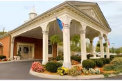 Covered front entrance of a brick nursing facility with columns, landscaping, and an American flag.