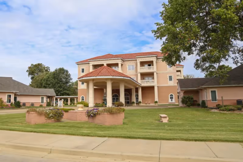 Exterior view of a peach-colored assisted living building with a circular entry porte-cochère and manicured lawn.