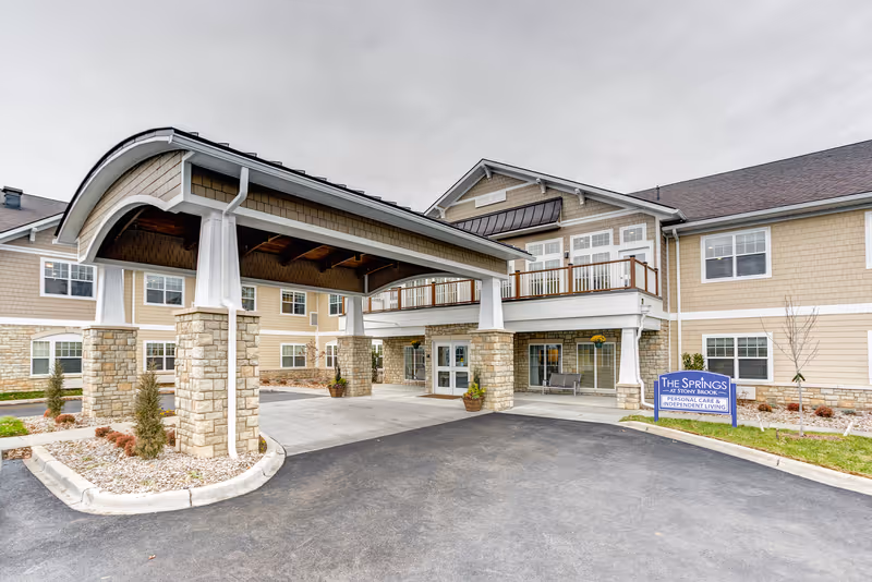 Exterior view of The Springs at Stony Brook senior living facility showing the main entrance with a covered driveway supported by stone pillars, beige siding, multiple windows, and a sign indicating personal care and independent living services.