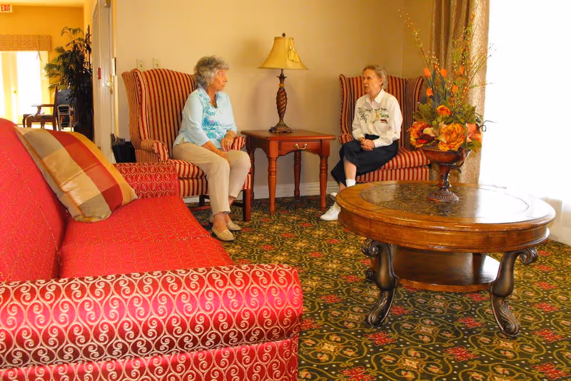 Two elderly women sitting and talking in a cozy living room area with patterned carpet, a red sofa with cushions, two striped armchairs, a wooden coffee table with a floral arrangement, and a side table with a lamp.