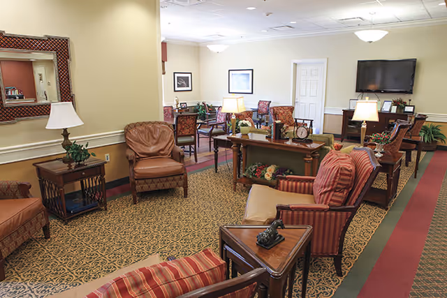 A cozy living room area in Richmond Place featuring multiple upholstered armchairs and sofas in warm tones, wooden side tables with lamps, a large patterned carpet, framed artwork on cream-colored walls, and a flat-screen TV mounted on the wall.