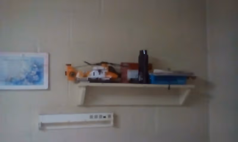 Wall shelf holds small items and a bottle; framed map on left, device below.