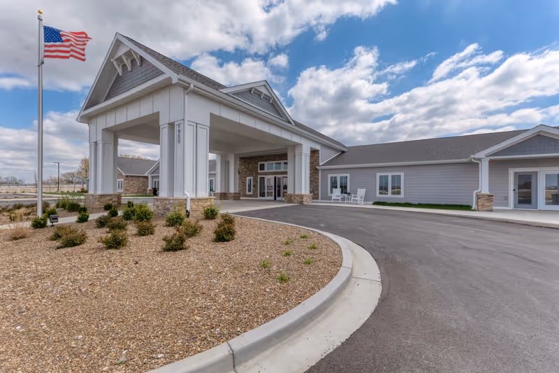 Front entrance of a single-story senior living facility with a covered porte-cochère, American flag, and landscaped circular driveway.