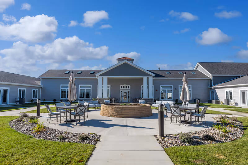 Outdoor patio area at Cedarhurst Senior Living of Nicholasville featuring round tables with chairs and closed umbrellas arranged around a circular stone fire pit, surrounded by landscaped grass and plants, with a single-story building in the background under a blue sky with scattered clouds.