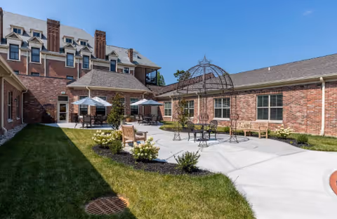 Sunlit brick courtyard with patio tables, benches, umbrellas, a decorative metal gazebo, and surrounding lawn.