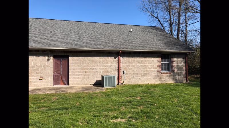 Exterior side of a single-story brick building with a door, window, rooftop, an outdoor HVAC unit, and a grassy yard under a clear sky.